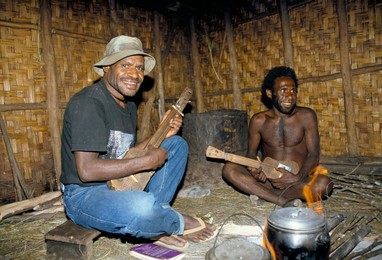 Dani men sit round fire, playing wooden guitar inside kitchen, South Beliam valley, Irian Jaya, Indonesia, Southeast Asia, Asia