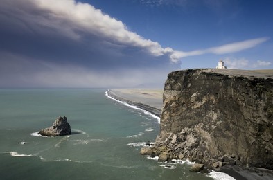 Headland with lighthouse at Dyrholaey looking towards a black volcanic sand beach stretching 15 miles into the distance and the ash plume of the Eyjafjallajokull eruption, near Vik i Myrdal, southern area, Iceland, Polar Regions
