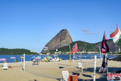 Rio de Janeiro, Brazil, A sandy beach with flagpoles displaying flags overlooks the serene seaside where umbrella canopies and people gather. In the background, mountains covered in greenery complete this peaceful travel destination.