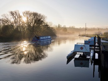 Row of pleasure boats at Radley Boathouse by the Thames, winter sunrise, with passing houseboat