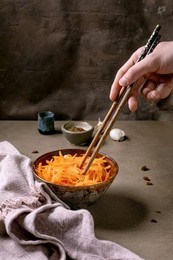Korean and Asian Cuisine. Man's Hand Uses Chopsticks To Pick Up Vegetable Salad of the rinated Carrots with Garlic from Ceramic Bowl. Seasoning In Small Bowls on Mocha Mousse Tones Table. Healthy Eating