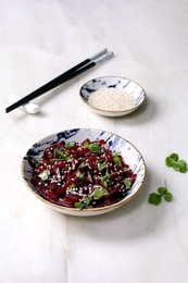 Korean-style Beet Salad. Marinated Grated Beetroot With Sesame Seeds and Coriander Leaves In Ceramic Bowl. Nearby Bowl With Sesame Seeds and Chopsticks On White Marble Table. Vegan Healthy Dish.