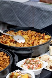 Appetizing Dish of Fried Potatoes with Corn Bell Peppers Mushrooms In Black Cooking Pan. Metal Spoon In Pan. Street Food with Side Dishes Visible In Foreground