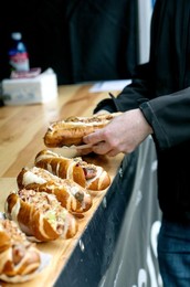 Person In Dark Jacket Selecting Sandwich From Wooden Counter. Fresh Filled Baguettes with Various Toppings Displayed Cafe Setting. Street Food Ready to Eat