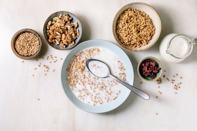 Top View of Sprouted Wheat Cereal with Milk Spoon Surrounded by Ingredients Nuts Seeds Granola Dried Cranberries. Healthy Breakfast Concept on Light Background